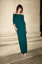 Woman wearing a teal off-shoulder dress standing against a beige wall.