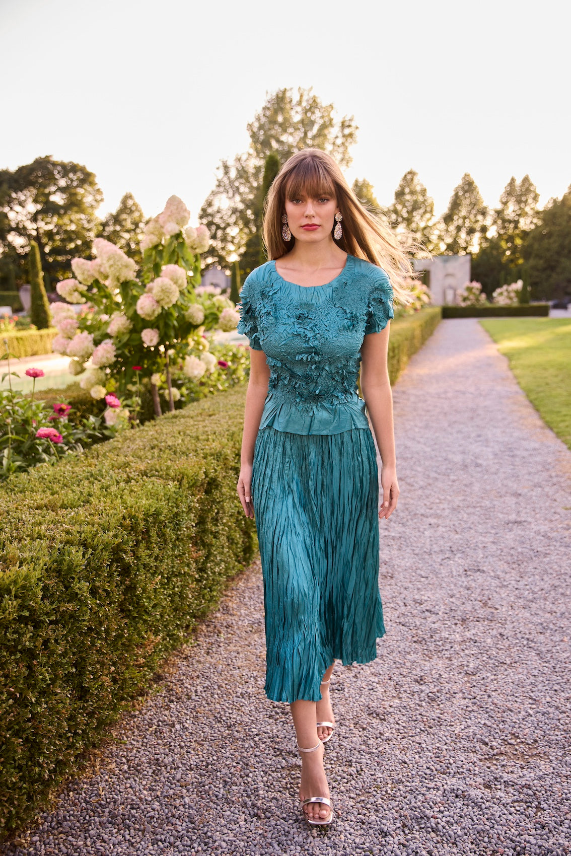 Woman in a teal dress walking along a garden path with flowers and trees in the background