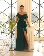 Woman in a dark green off-shoulder evening gown with a high slit standing indoors.