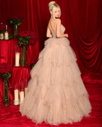 Woman in a beige ball gown standing in front of red curtains with floral decorations.