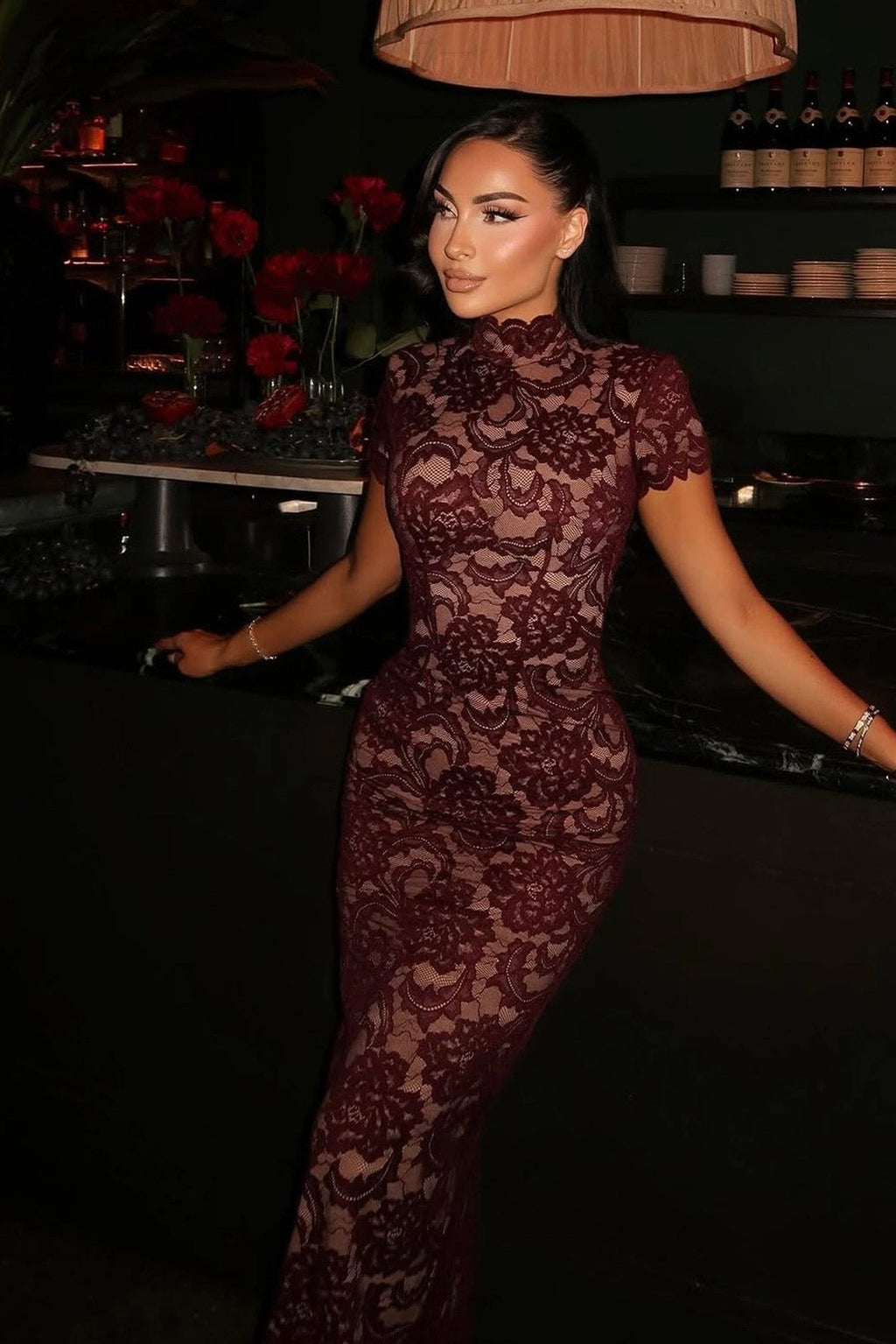 Woman in a burgundy lace dress standing in a dimly lit room with red flowers and a lamp.