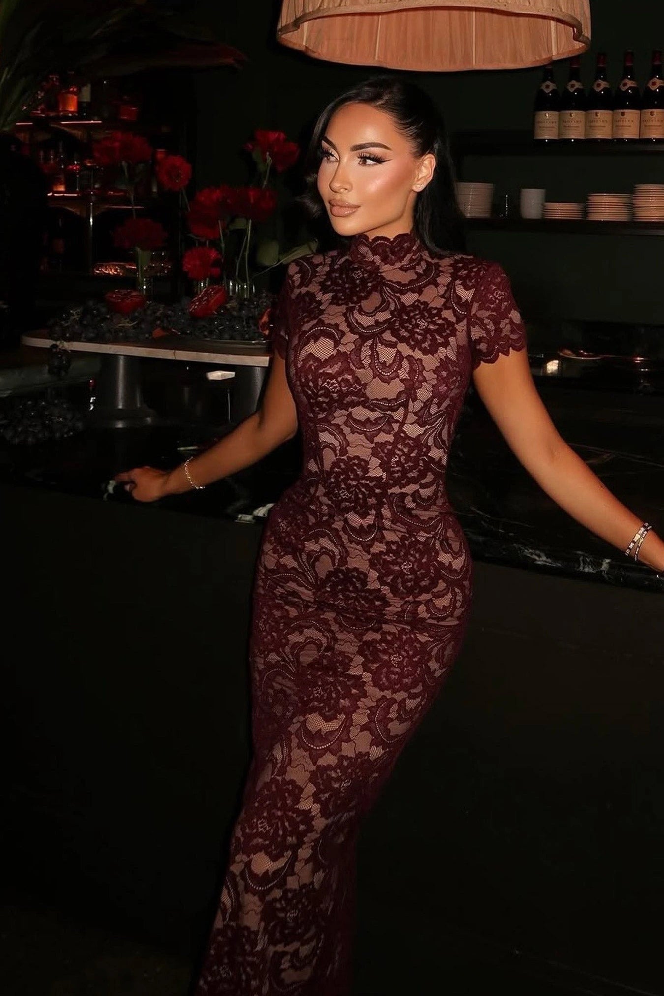 Woman in a burgundy lace dress standing in a dimly lit room with red flowers and a lamp.