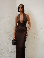 Woman in a brown dress holding a black handbag against a textured white wall.