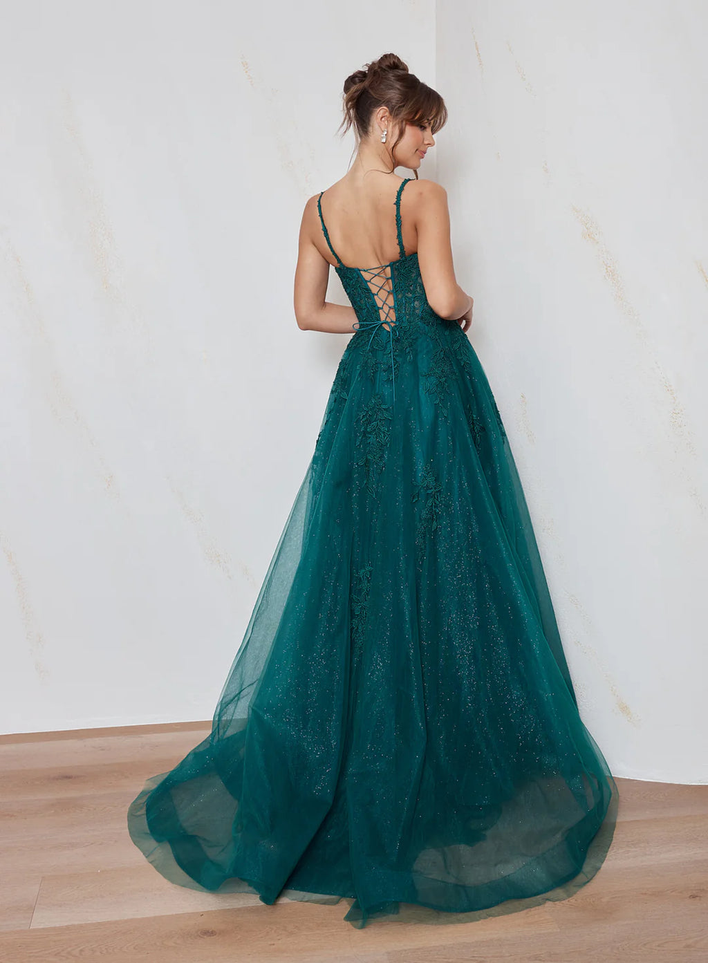 Woman wearing a teal evening gown with a lace-up back against a white wall.