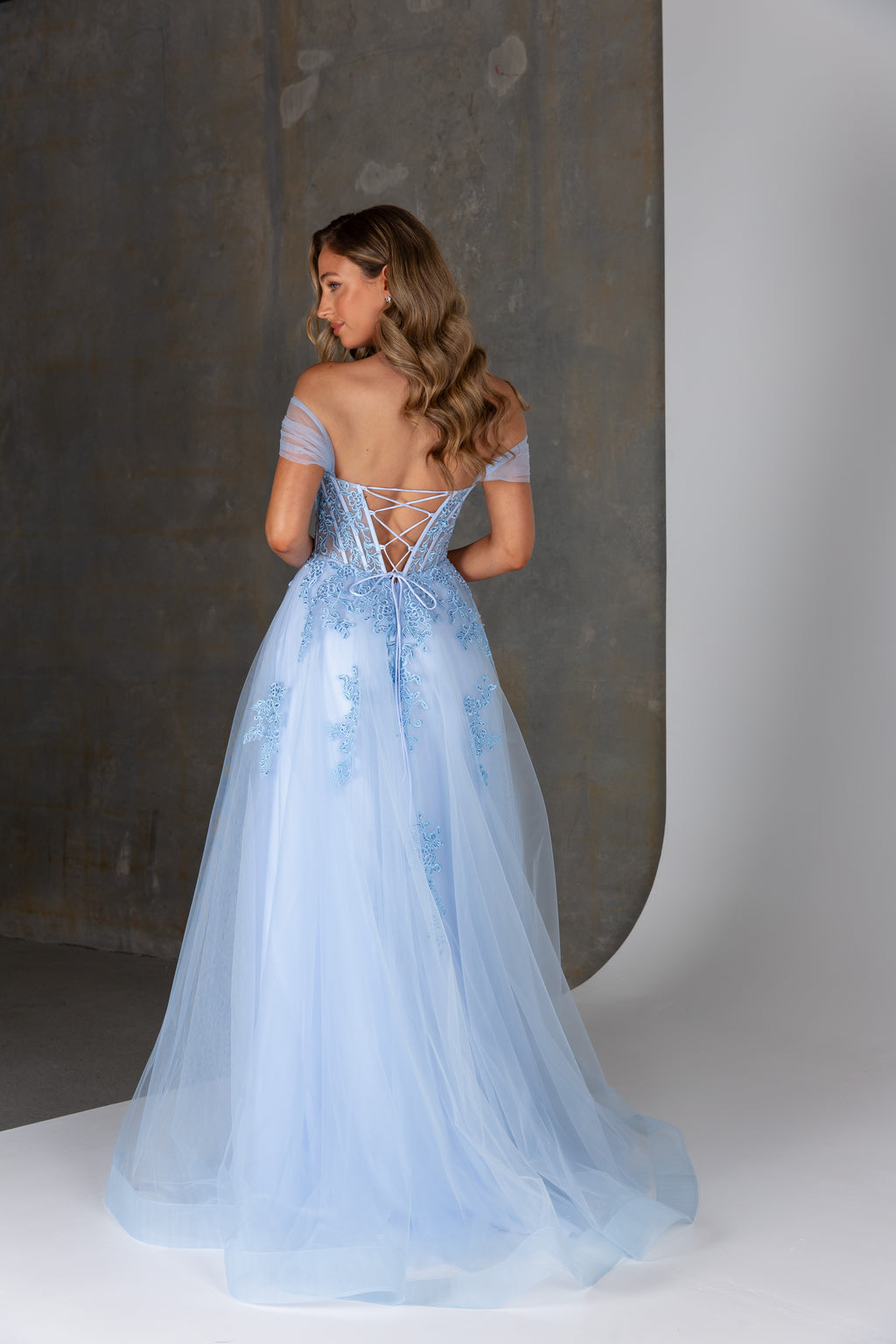 Woman wearing a light blue evening gown with a lace-up back against a neutral background