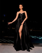 Woman in a black strapless gown with a high slit standing against a dark background.