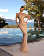 Woman in a beige evening gown standing by a pool with a scenic background