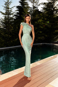 Woman in a light green one-shoulder dress standing by a pool with trees in the background