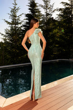 Woman in a light green one-shoulder dress standing by a pool with trees in the background