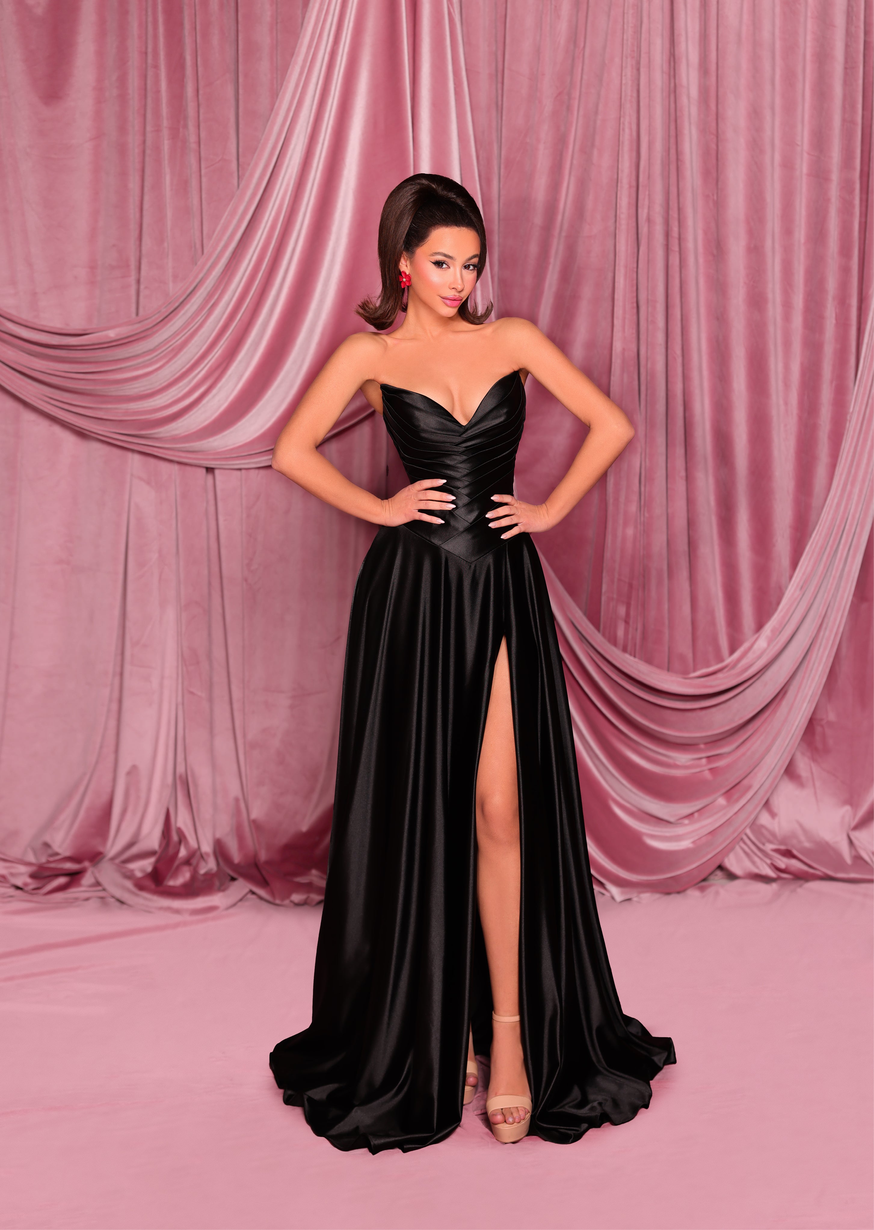 Woman in a black evening gown with a high slit standing against a pink draped curtain backdrop.