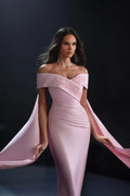 A woman models a floor-length off-the-shoulder evening gown in soft blush pink. The dress features a structured sweetheart neckline with lace applique, a fitted ruched bodice, and dramatic flowing cape-like sleeves that she holds out to both sides against a dark studio background.