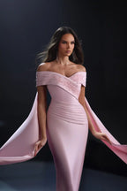 A woman models a floor-length off-the-shoulder evening gown in soft blush pink. The dress features a structured sweetheart neckline with lace applique, a fitted ruched bodice, and dramatic flowing cape-like sleeves that she holds out to both sides against a dark studio background.