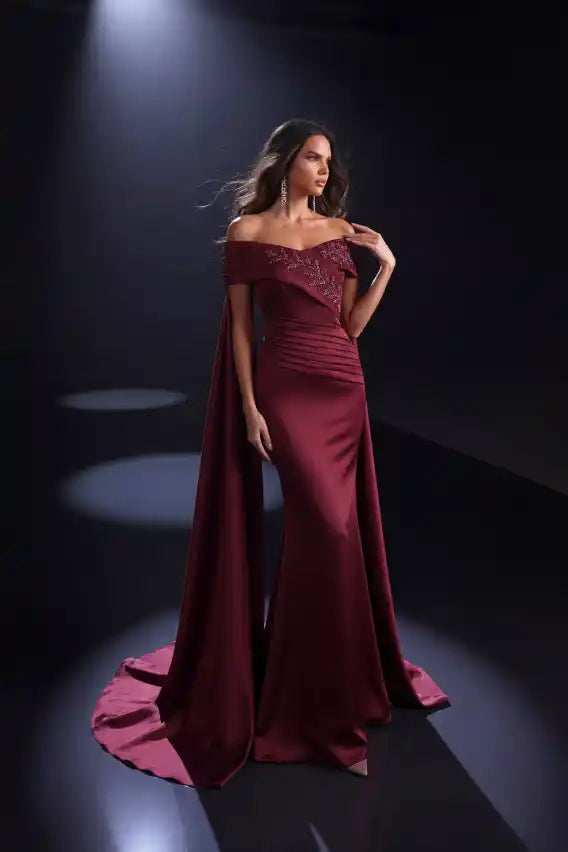 A woman models an elegant off-the-shoulder evening gown in deep burgundy with a long flowing cape train, set against a dark stage background with dramatic spotlights.