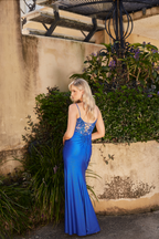 Woman in a blue evening gown standing against a textured wall with plants.