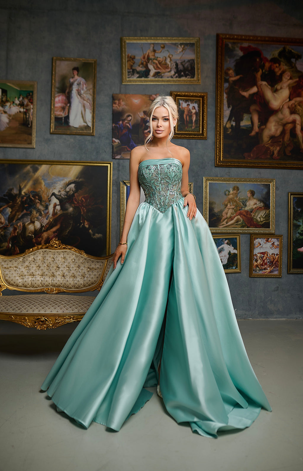 Woman in a teal strapless gown standing in a room with classical paintings on the wall.