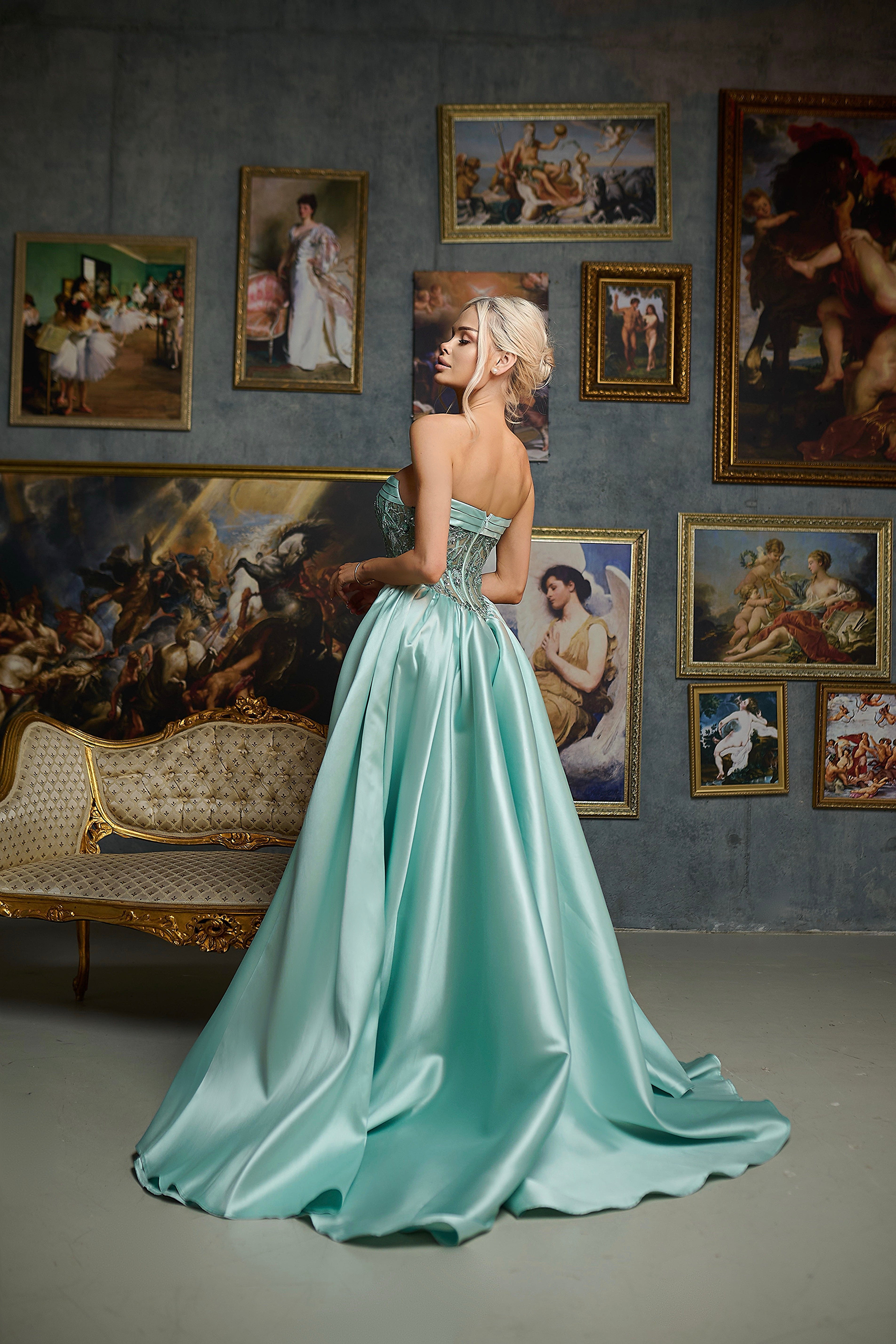Woman in a light blue gown standing in a room with classical paintings on the wall.